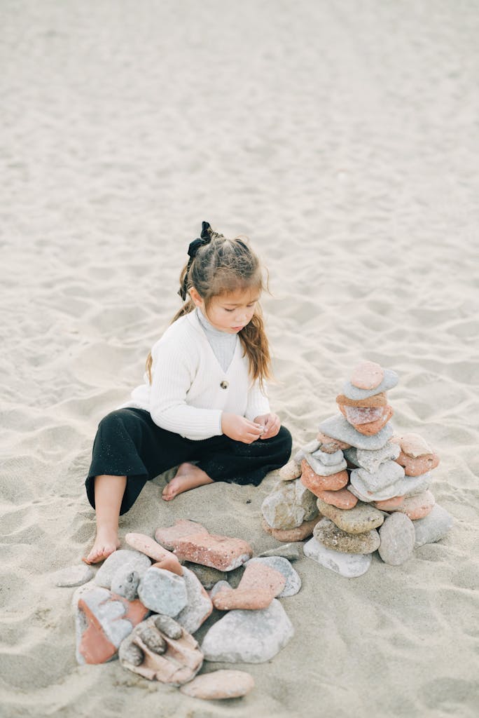 A young girl plays by stacking rocks on a sandy beach, enjoying a peaceful day outdoors.
